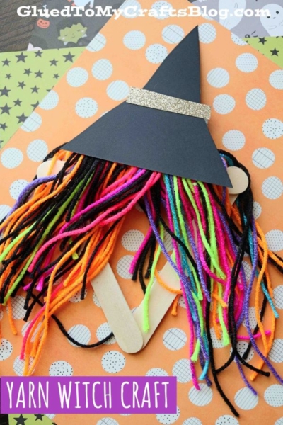 Colorful yarn strands form witch hair under a black paper hat with a gold band, attached to craft sticks, placed on orange patterned paper—perfect for your next yarn witch craft project. Text: Yarn Witch Craft.