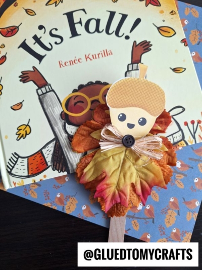 A children’s book titled It’s Fall! by Renée Kurilla lies on a table. In front is a fall craft inspired by acorn friend puppets, with a popsicle stick, fake autumn leaves, paper acorn face, button, and raffia. @GLUEDTOMYCRAFTS is labeled below.