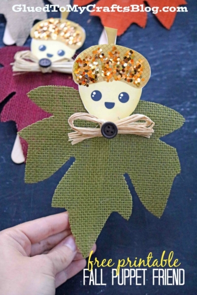 A hand holds a green, fabric leaf puppet with a smiling acorn face, gold sequin cap, and a raffia bow with a button—one of the adorable acorn friend puppets. Another burgundy puppet is in the background. Text reads free printable FALL PUPPET FRIEND.