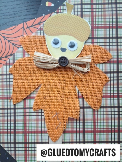 A craft project featuring acorn friend puppets with acorn-shaped faces, googly eyes, and smiles, plus a raffia bow and button on an orange burlap leaf—all set on a plaid, Halloween-themed background. Text reads @GLUEDTOMYCRAFTS.
