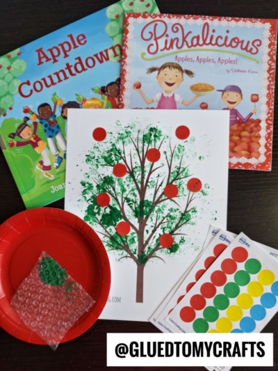 Children’s books “Apple Countdown” and “Pinkalicious: Apples, Apples, Apples!” sit beside a bubble wrap stamped apple tree craft with apple stickers, a red plate, and sticker sheets. Text reads “@GLUEDTOMYCRAFTS.”.