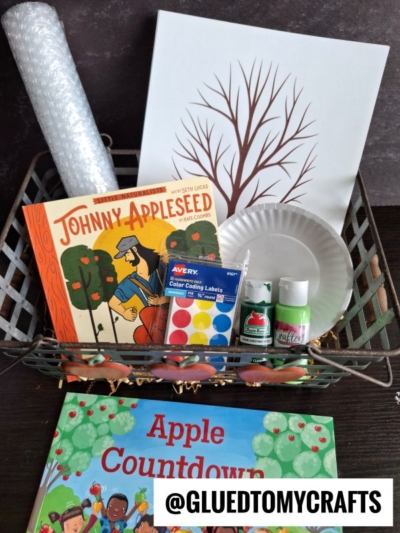 A basket contains a Johnny Appleseed book, an Apple Countdown book, paint, paper plates, color labels, bubble wrap for a bubble wrap stamped apple tree craft, a roll of plastic, and a canvas with a tree outline. A label says @GLUEDTOMYCRAFTS.