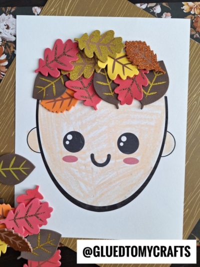 A mixed media leaf hair drawing of a smiling face features foam autumn leaves arranged on top as hair, with extra foam leaves scattered nearby. The handle @GLUEDTOMYCRAFTS appears at the bottom.