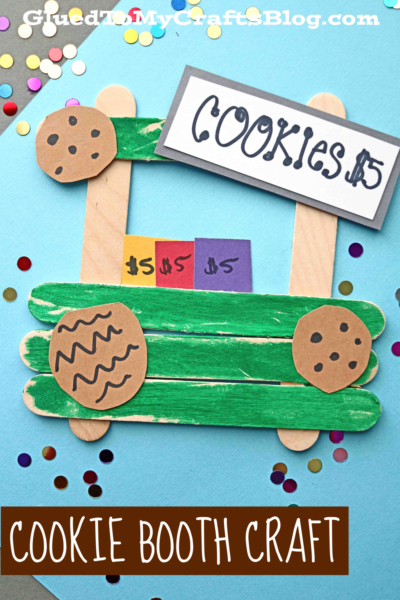 A handmade popsicle stick Girl Scout cookie booth craft features green-painted sticks, paper cookies, colorful price tags, and a COOKIES $5 sign on a blue background with scattered confetti.