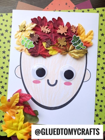 A children’s craft shows a smiling face outline with large googly eyes and red cheeks. The mixed media leaf hair features colorful artificial leaves and glittery foam shapes. The background is pale green with black stars.