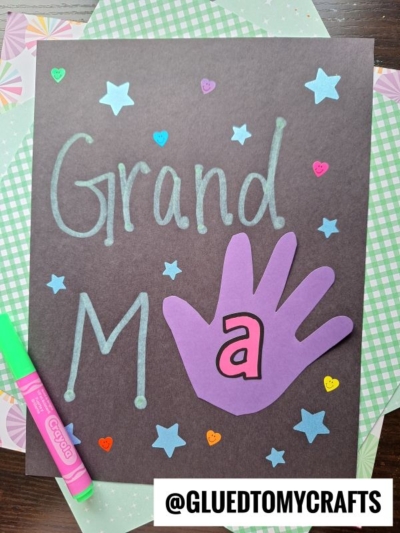 Handmade card on black paper with Grand M a written in blue marker, a purple paper hand cutout with an a, star and heart stickers—perfect for celebrating National Grandparent’s Day—and a green marker nearby. Username @GLUEDTOMYCRAFTS at the bottom.