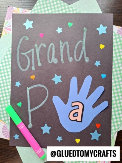 A craft project for National Grandparent’s Day features Grandpa spelled with a handprint on black paper, decorated with stars and hearts. Two markers and patterned papers are in the background. @GLUEDTOMYCRAFTS text appears at the bottom.