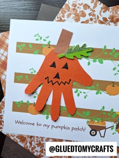 A handmade pumpkin craft using an orange handprint cutout with a jack-o-lantern face, green leaf, and brown stem is featured on a cheerful “Welcome to my pumpkin patch!” card. Plaid papers create a charming background for this festive design.