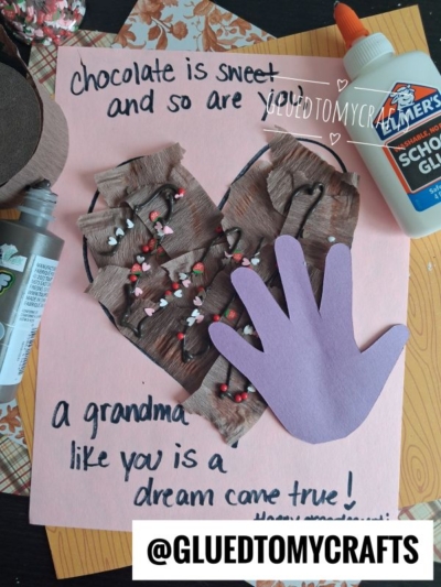 A handmade card with a brown heart decorated like chocolate, a purple paper hand cutout, and a message about grandma—perfect for celebrating National Grandparent’s Day. Glue, craft paint, and scissors are nearby. @GLUEDTOMYCRAFTS is at the bottom.