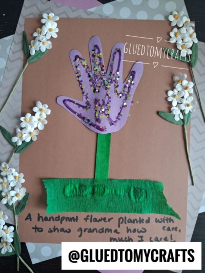 A handmade card with a purple handprint flower, decorated with confetti, green crepe paper stem, faux flowers, and the message: A handprint flower planted with love to show grandma how much I care! Perfect for celebrating National Grandparent’s Day.