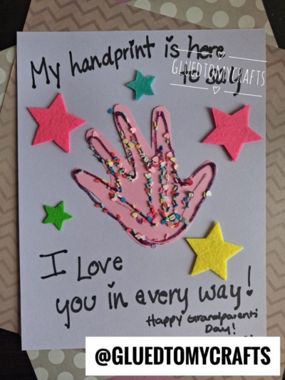 A handmade card with a pink paper handprint decorated with sprinkles, surrounded by colorful felt stars. The card says, My handprint is here! I love you in every way! Happy Grandparents Day! Celebrate National Grandparent’s Day with this heartfelt keepsake.