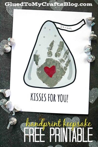 A printed Hersheys Kiss outline features a child’s gray handprint and a red heart, labeled KISSES FOR YOU! and Jayden, creating a sweet handprint kisses keepsake on a black surface with silver heart decorations.
