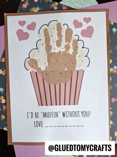 A handmade card features a cupcake with a brown handprint as the muffin top, pink hearts above, and the text: I’d be muffin without you! Love ______. Perfect for National Grandparent’s Day, the card is surrounded by floral and purple paper.