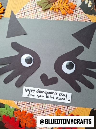 A handmade cat face craft with googly eyes, black paper cutouts, and a note saying Happy Grandparents Day from your little rascal—perfect for celebrating National Grandparent’s Day. Autumn leaves and plaid paper are in the background.