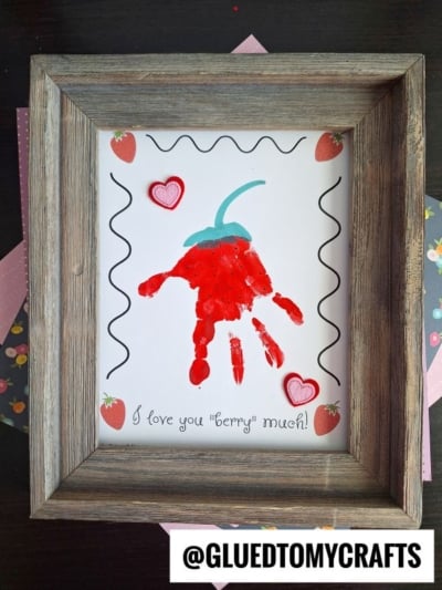 A framed craft features a red handprint strawberry with a green stem, surrounded by drawn hearts and strawberries, and the words I love you berry much! at the bottom—perfect for celebrating National Grandparent’s Day.