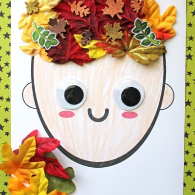 A paper craft of a smiling face with large googly eyes, red cheeks, and colorful autumn leaves for mixed media leaf hair. The image has a “Leaf Hair Craft” label and a website name at the top.