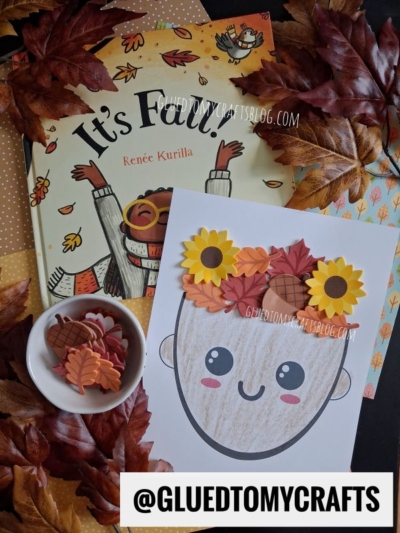 A fall craft scene with a children's book, fake leaves, a bowl of autumn-themed cutouts, and a smiling face drawing decorated with mixed media leaf hair and sunflowers, with @GLUEDTOMYCRAFTS at the bottom.