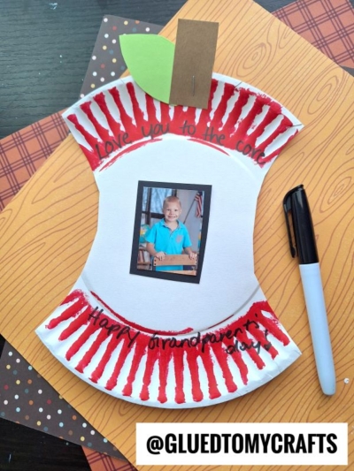 Apple core craft with a child’s photo in the center, red painted edges, green paper leaf, and handwritten messages: Love you to the core and Happy National Grandparent’s Day! Nearby are a marker and patterned papers.