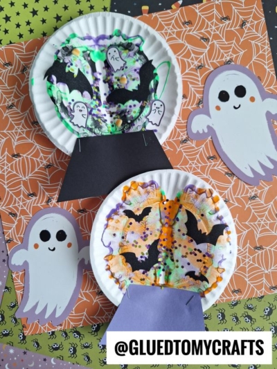 Two Halloween-themed craft projects resembling snow globes feature paint splat backgrounds made with paper plates, coffee filters, and cut-outs of bats and ghosts—all set on colorful Halloween-patterned paper. Text reads @GLUETOMYCRAFTS.