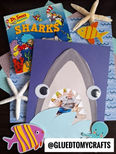 A shark mouth picture frame craft featuring a child’s photo inside a paper shark mouth, large googly eyes, colorful sea animal cutouts on ocean-patterned paper, displayed beside a Dr. Seuss “Sharks” book and playful starfish.