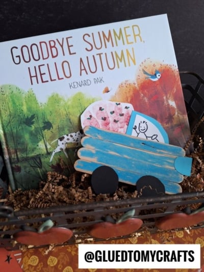 A children’s book titled Goodbye Summer, Hello Autumn by Kenard Pak is displayed in a basket with brown paper shreds, alongside a blue popsicle stick farm truck craft carrying a heart.