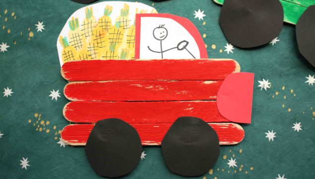 A popsicle stick farm truck made of red sticks, black paper wheels, and a red paper heart bumper. The truck carries yellow corn and has a stick figure driver drawn in the cab, perfect for playful farm-themed crafts.