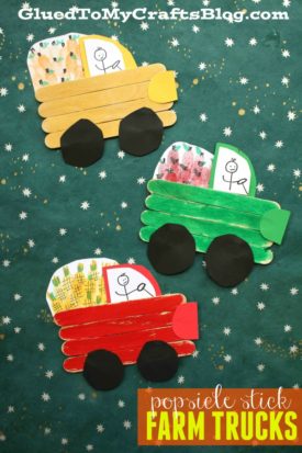 Three colorful popsicle stick farm trucks—yellow, green, and red—with black paper wheels and hand-drawn stick figures inside are displayed on a starry green background.