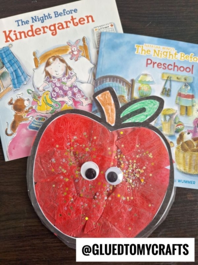 Two children’s books titled The Night Before Kindergarten and The Night Before Preschool are displayed behind a red stained glass apple craft with googly eyes and glitter, on a dark surface. @GLUEDTOMYCRAFTS appears at the bottom.