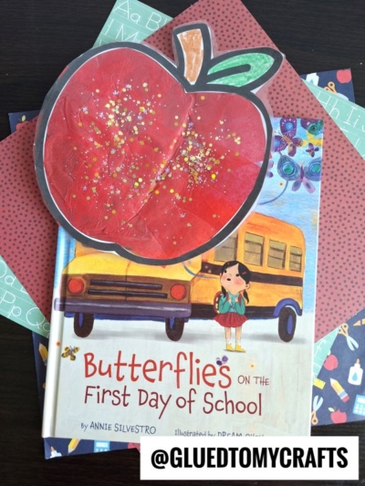 A children’s book titled Butterflies on the First Day of School is topped with a large stained glass apple on a background of colorful paper sheets. The @GLUEDTOMYCRAFTS watermark appears at the bottom.