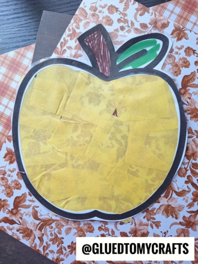 A stained glass apple craft featuring yellow tissue pieces inside a black outline, set on patterned autumn-themed paper. It has a brown stem, green leaf, and @GLUEDTOMYCRAFTS labeled at the bottom.