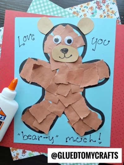 A blue card with a bear made of torn brown paper, googly eyes, and a drawn smile. It says Love you bear-y much!—the perfect craft for National Grandparent’s Day. A bottle of glue and patterned paper are nearby.