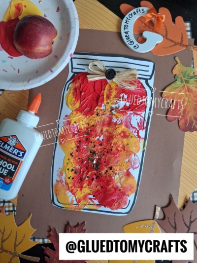 A fall craft project shows an apple stamped mason jar jam design made from a paper jar decorated with red, yellow, and orange paint, glitter, and a raffia bow. Elmer’s glue, a paper plate with an apple, and autumn leaf cutouts are nearby. @GLUETOMYCRAFTS appears on the image.