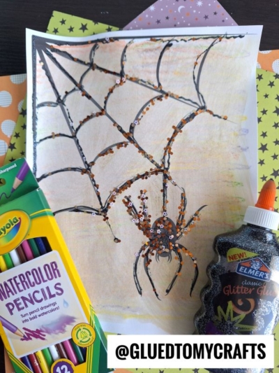 A drawing of a black spider on an orange web sparkles with glitter, making a perfect spider web craft for kids. Crayola watercolor pencils and Elmer’s glitter glue sit nearby, while patterned orange, black, and purple papers are underneath.