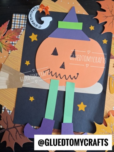 A paper craft of a Flying Pumpkin Witch—with green legs, a purple hat, and boots—sits on a broomstick, surrounded by stars and autumn leaves on a black background. @GLUETOMYCRAFTS is written at the bottom.