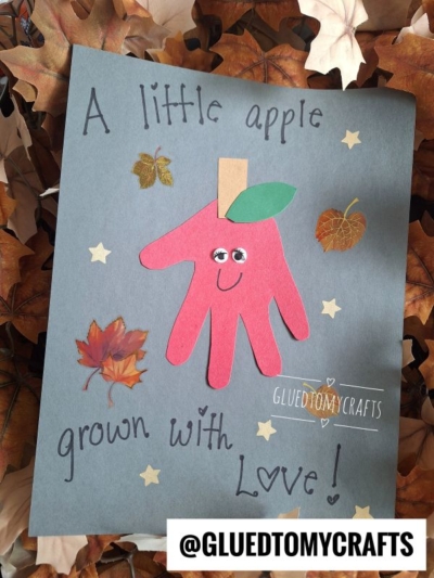 A handmade craft features a red handprint cutout decorated as an apple with googly eyes and a smile, surrounded by leaf stickers on dark paper—a charming choice for fall keepsake ideas. Text reads, “A little apple grown with love!”.