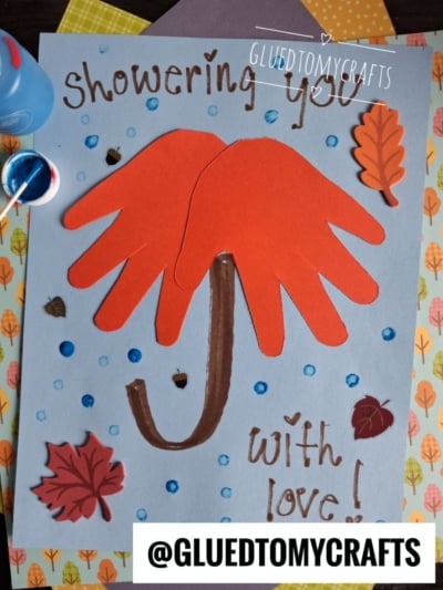 Children’s craft with two red handprints forming an umbrella, a brown handle, leaves, and blue dots on light blue paper. Text says “Showering you with love!” and @GLUEDTOMYCRAFTS. A charming addition to your fall keepsake ideas collection!.