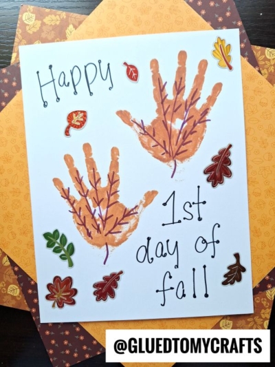 A fall-themed craft with two handprints resembling turkeys, leaf stickers, and the words “Happy 1st day of fall!” on white paper—perfect for those searching for fall keepsake ideas. Decorative autumn papers are layered underneath to complete this charming seasonal project.