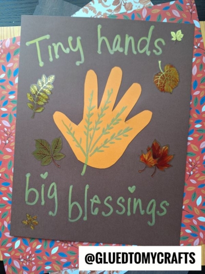 A brown card with a cutout of a hand in orange paper, decorated with leaf stickers—a perfect addition to your fall keepsake ideas. Green text reads Tiny hands big blessings. @GLUEDTOMYCRAFTS is printed on the bottom.