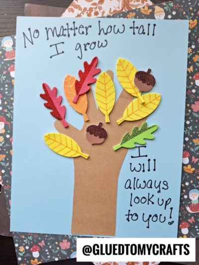 A handprint tree craft with felt leaves and acorns on blue paper, perfect for those looking for fall keepsake ideas. Text reads, No matter how tall I grow, I will always look up to you! @GLUEDTOMYCRAFTS is at the bottom.