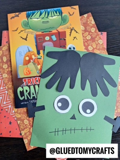 A Halloween craft scene with a Frankenstein-themed book, orange patterned papers, and a green handprint Frankenstein face made from paper with black hair, big eyes, and stitched mouth. @GLUEDTOMYCRAFTS is at the bottom.