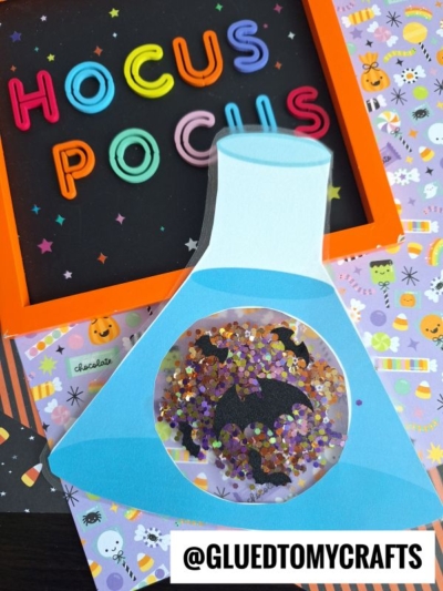 A colorful Hocus Pocus sign and a Halloween-themed card shaped like a stained glass halloween beaker, filled with sparkly confetti and featuring a bat silhouette. @GLUEDTOMYCRAFTS is written at the bottom.