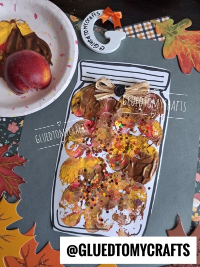 A paper jar craft inspired by apple stamped mason jar jam, featuring brown and orange paint, glitter, a bow, and a button, sits on a table with fake leaves, a peach, and a paintbrush. The handle says GLUEDTOMYCRAFTS. The username appears at the bottom.