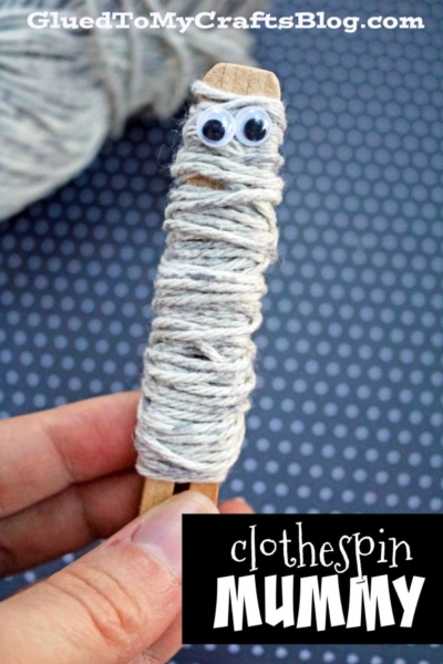 A hand holds a wooden clothespin wrapped in white string with two googly eyes on top, resembling a mummy. Perfect for clothespin crafts for kids, the dark background with small white dots sets off this spooky clothespin craft.