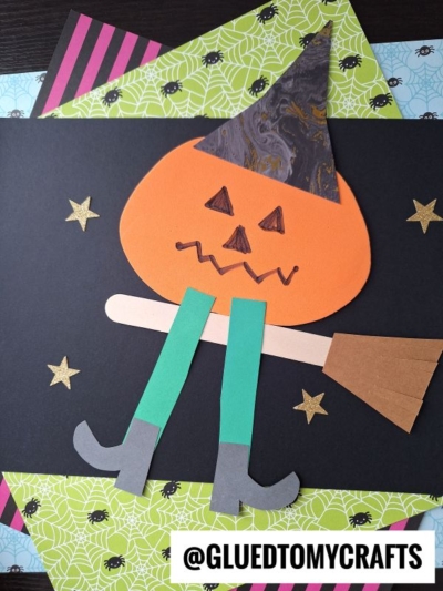 A Halloween craft featuring a Flying Pumpkin Witch with a paper pumpkin, witch hat, broom, green legs, and gray boots, set on black paper with gold stars and colorful spider web-themed papers. @GluedToMyCrafts is visible.