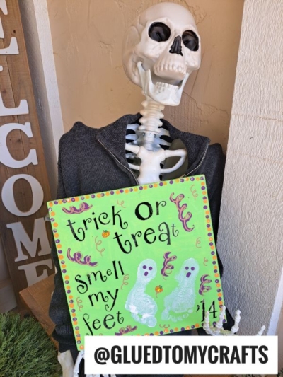 A skeleton in a hoodie holds a green sign that says trick or treat, smell my feet with painted ghosts and colorful decorations—making it a fun addition to your collection of fall keepsake ideas. The sign includes the Instagram handle @GLUEDTOMYCRAFTS.