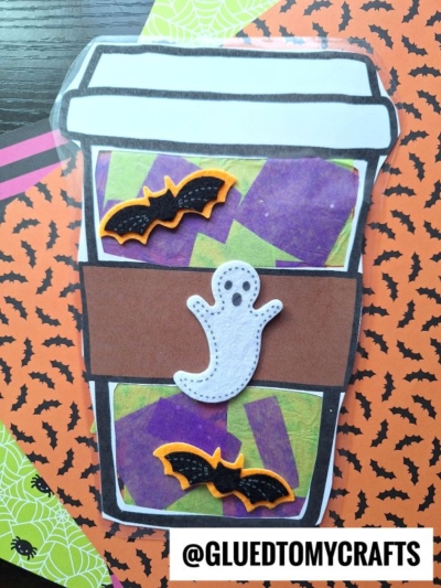 A Halloween-themed paper coffee cup craft decorated with two black bats, a white ghost, and colorful purple and green shapes captures the cozy spirit of autumn. The background features orange paper with spiderweb patterns, evoking the warmth of a stained glass pumpkin spice latte on a crisp fall day.