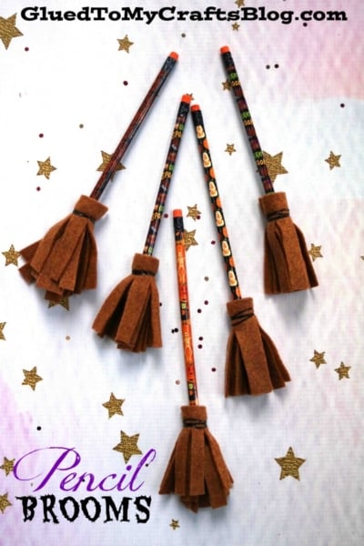 Five Halloween-themed pencils are decorated with brown felt to resemble pencil witch brooms, arranged on a light background with scattered gold star confetti. Pencil Brooms is written at the bottom.