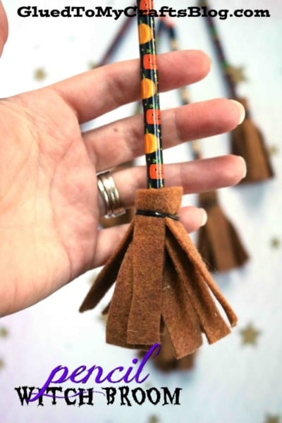 A hand holds a pencil decorated with a pumpkin pattern, transformed into one of the adorable pencil witch brooms using brown felt fringed at the end. The text on the image reads, pencil WITCH BROOM.