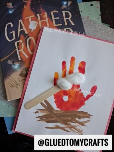 Children’s art project showing a handprint campfire made with red, yellow, and orange paint, topped with cotton balls as marshmallows on a craft stick—perfect for fall keepsake ideas. Book and papers are in the background.