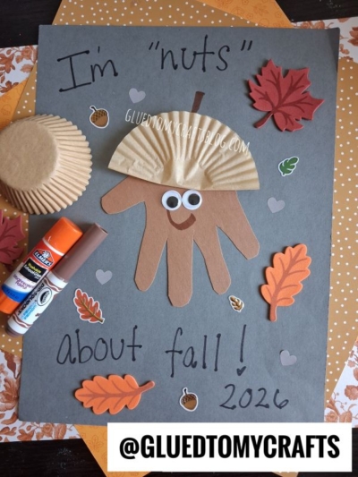 A fall craft features a handprint squirrel with a cupcake liner hat, googly eyes, and paper leaves on gray paper. Text says "I'm nuts about fall! 2026." Glue, markers, and liners are beside it—perfect inspiration for anyone looking for fun fall keepsake ideas.